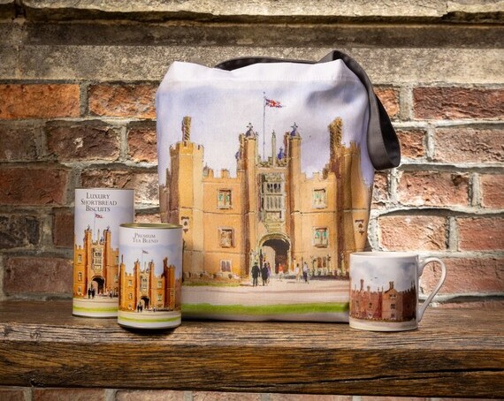 A tote bag, mug and food gifts against a red Tudor-brick wall printed with a watercolour image of Hampton Court Palace, 