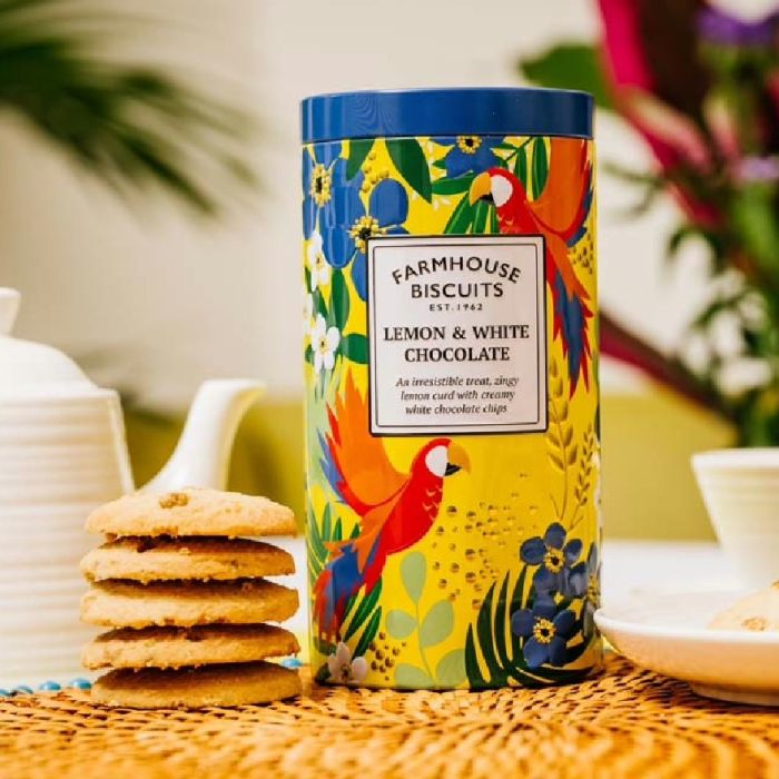 A colourful tin of lemon and white chocolate biscuits with cookies, a teapot, cup, and flowers arranged on a table.