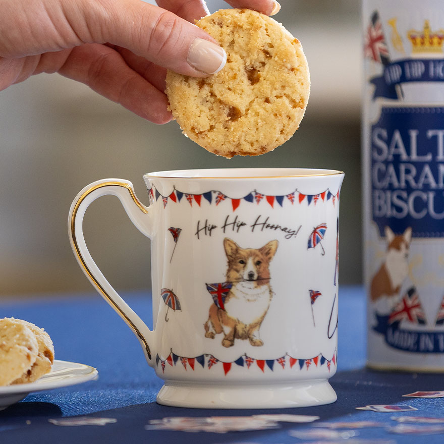 A close-up of a hand dunking a biscuit into a mug of tea. The mug is printed with a corgi dog holding a Union Jack.
