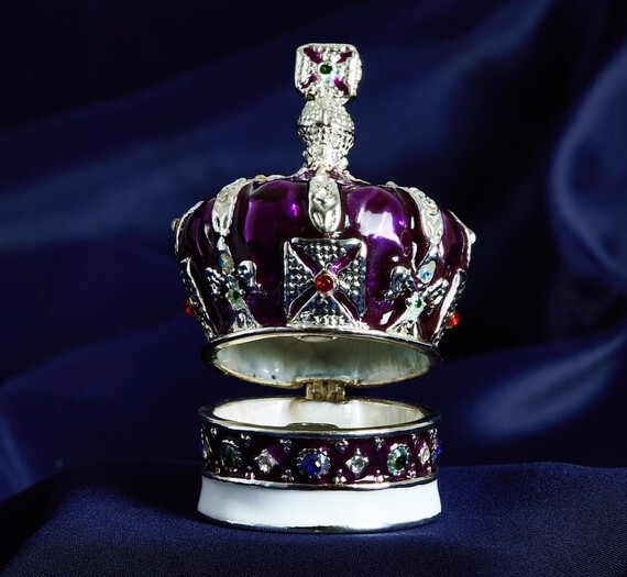 A trinket tin in the shape of a royal crown in purple and silver, slightly hinged open.