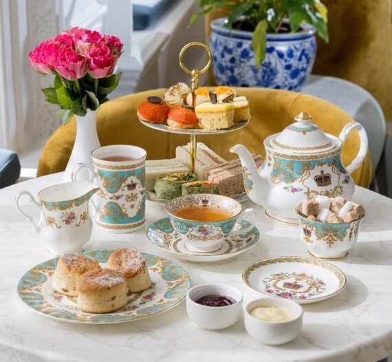 A set of fine china in white, aqua and gold laid out on a round table with scones, sandwiches and tea.