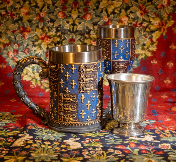 A tankard and two drinking goblets in a medieval style against a red and green tapestry.